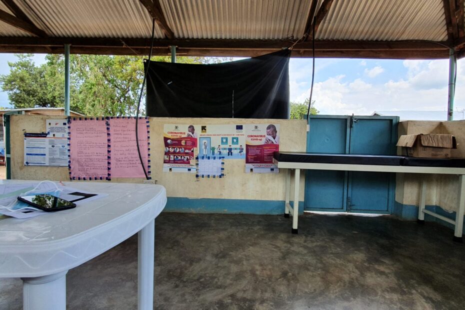 Image of a white desk in the foreground, with papers and a face mask on it, with colorful walls in the background, on which are hanging posters with text about COVID-19. Blue skies, clouds, and trees are visible between the wall partitions and the ceiling, and a few people are visible sitting outside the room.