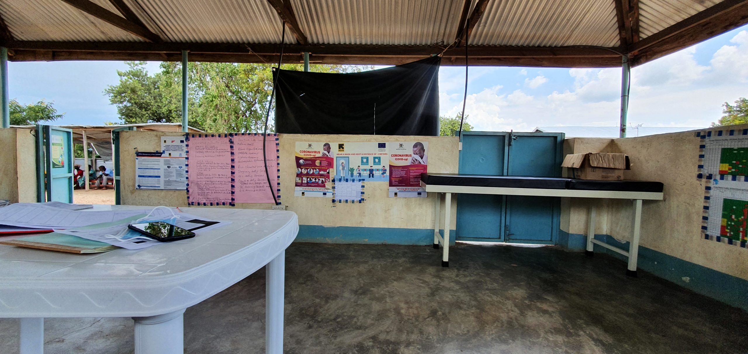 Image of a white desk in the foreground, with papers and a face mask on it, with colorful walls in the background, on which are hanging posters with text about COVID-19. Blue skies, clouds, and trees are visible between the wall partitions and the ceiling, and a few people are visible sitting outside the room.