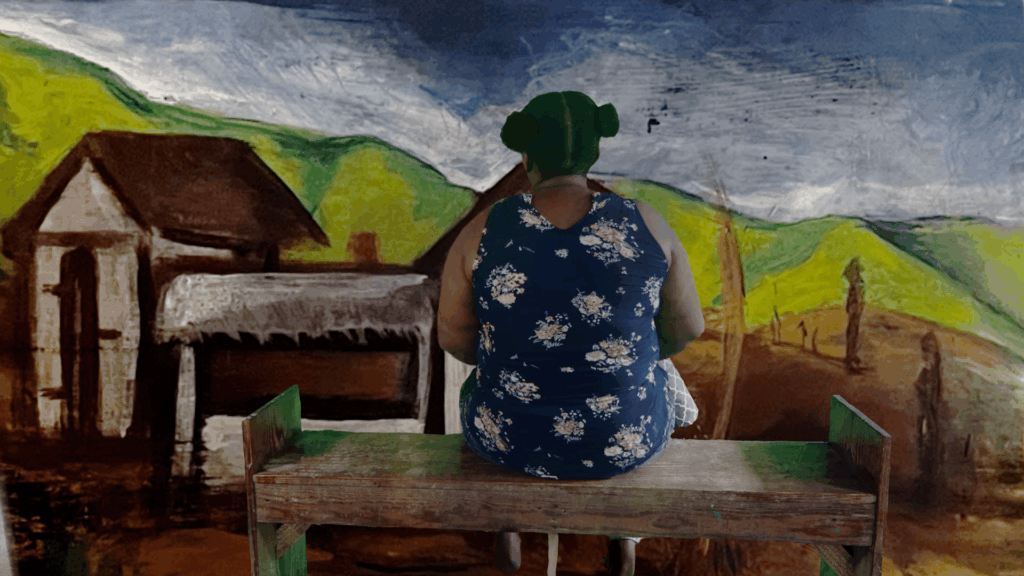 A woman sitting at a bench facing a mural of a colorful landscape with her back to the viewer.