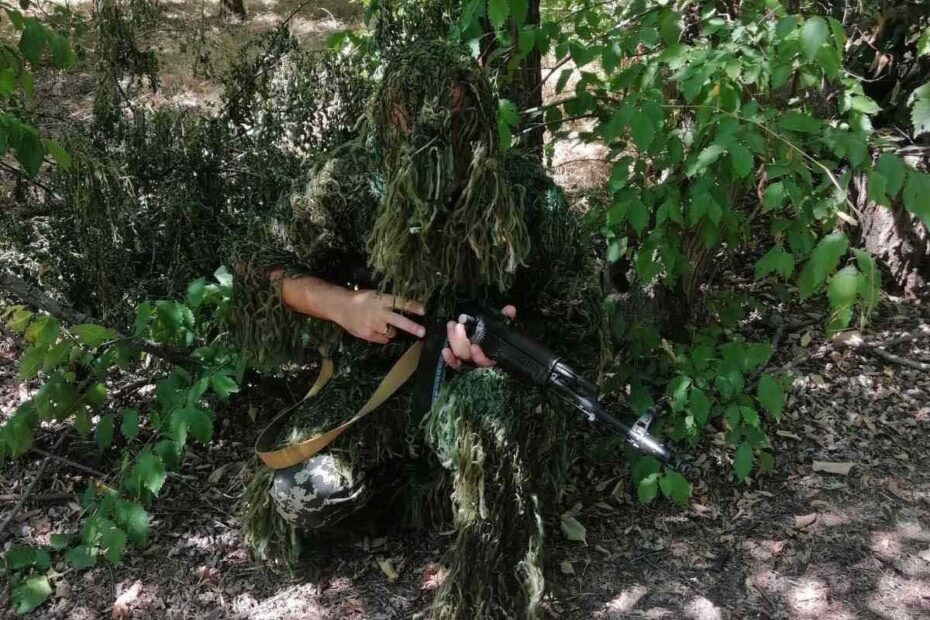 A soldier, covered by a camouflage net, crouches next to a tree and greenery.