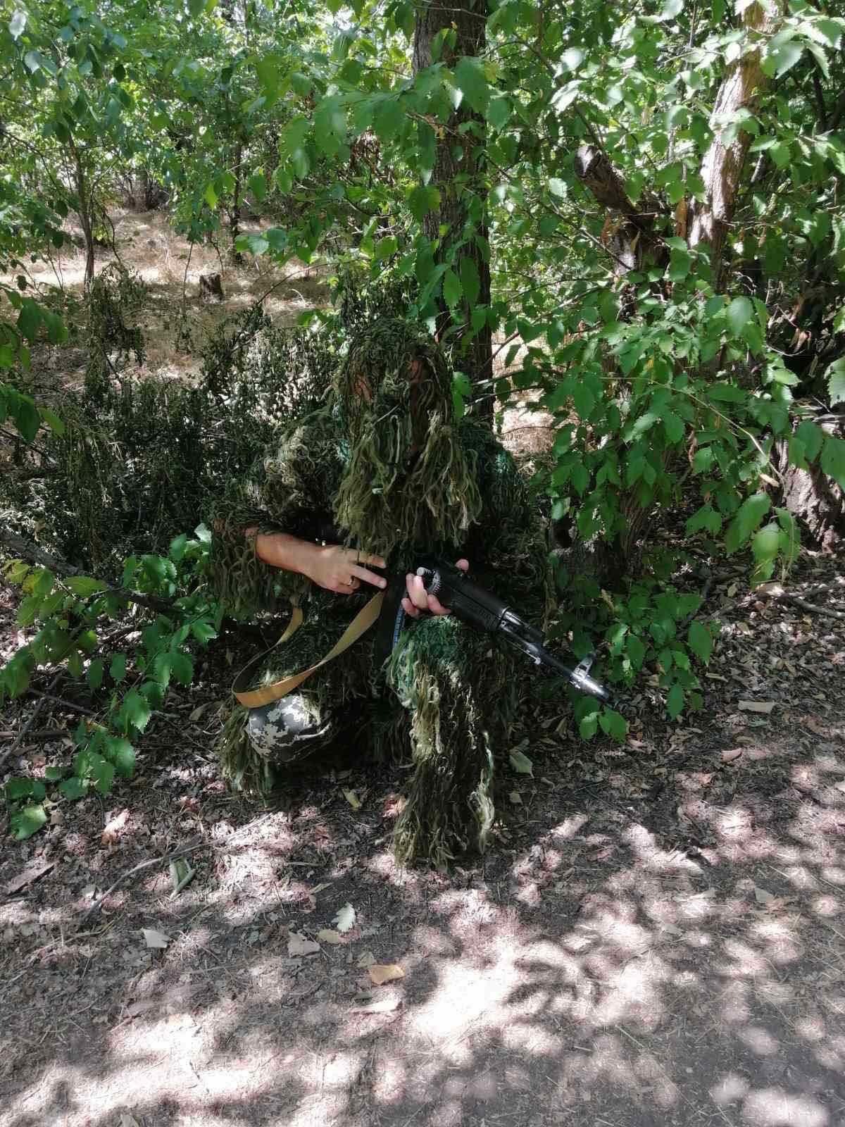 A soldier, covered by a camouflage net, crouches next to a tree and greenery.