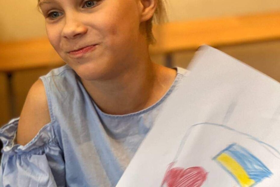 A girl, looking away from the camera, holds up a piece of paper with a drawing of a heart and a Ukrainian flag.